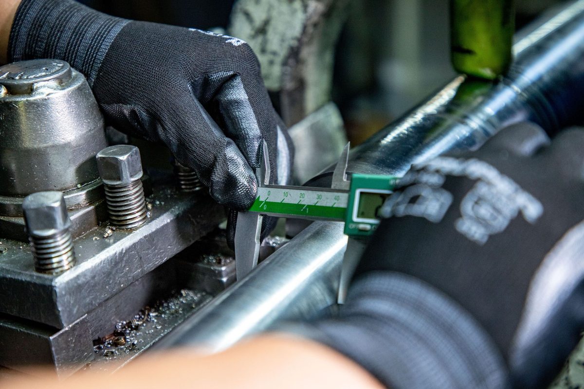 Primo piano di una mano guantata che misura con un calibro digitale un pezzo metallico su un tornio, a illustrare la verifica di un pezzo prima dell'acquisto.
